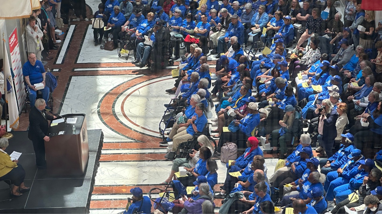 a photo taken from above of hundreds of people in blue shirts listen to a speaker in the Great Hall at the Massachusetts State House