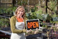 Small business owner, mature woman (50s), holding OPEN sign in plant nursery.