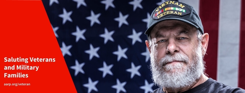 Photo of  male with a white beard wearing a Vietnam Veteran cap standing in front of an American flag.