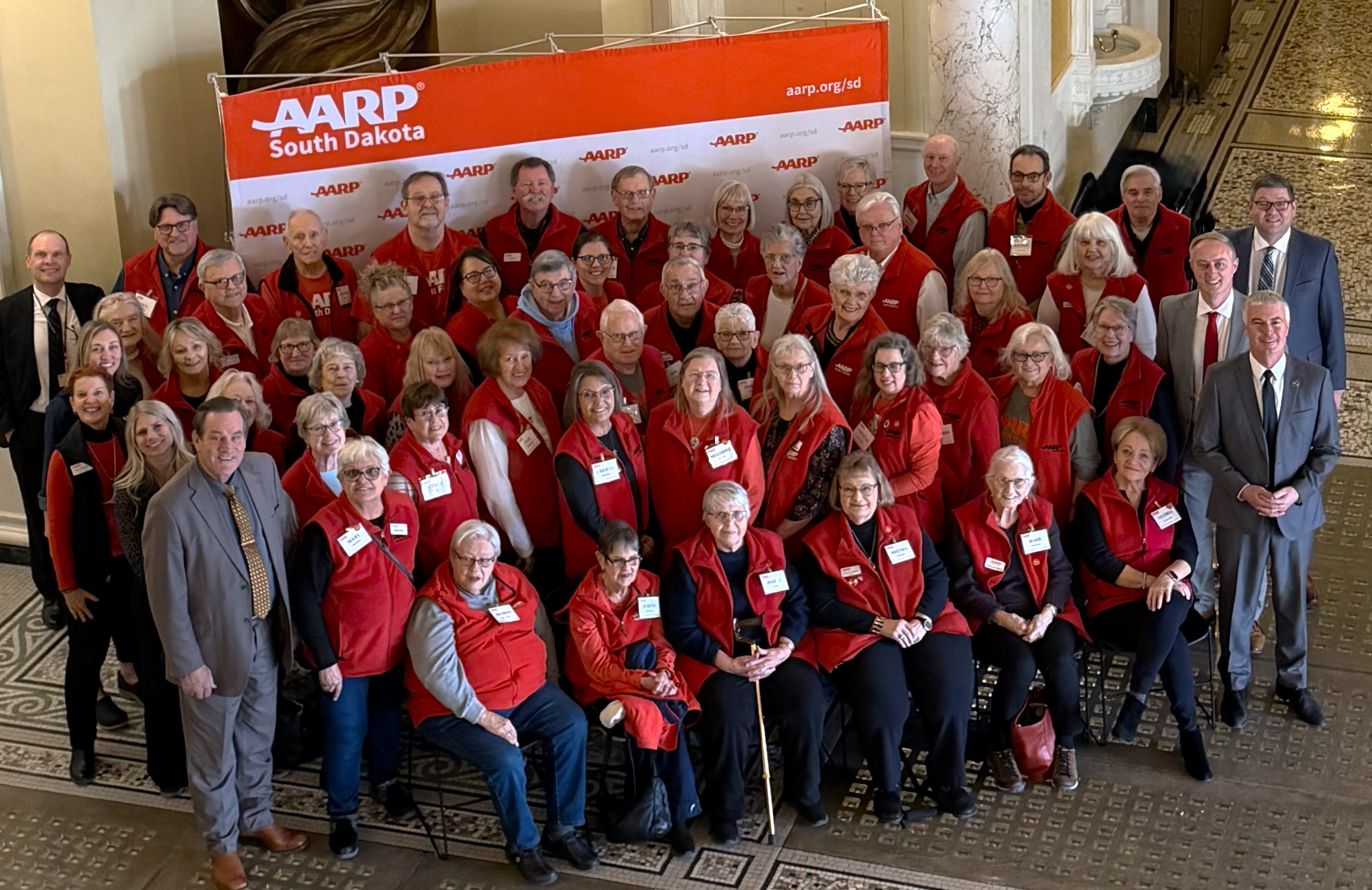 A group of men and women in red vests representing AARP South Dakota at the State Capitol pictured with the Governor, Lt. Governor and Attorney General.