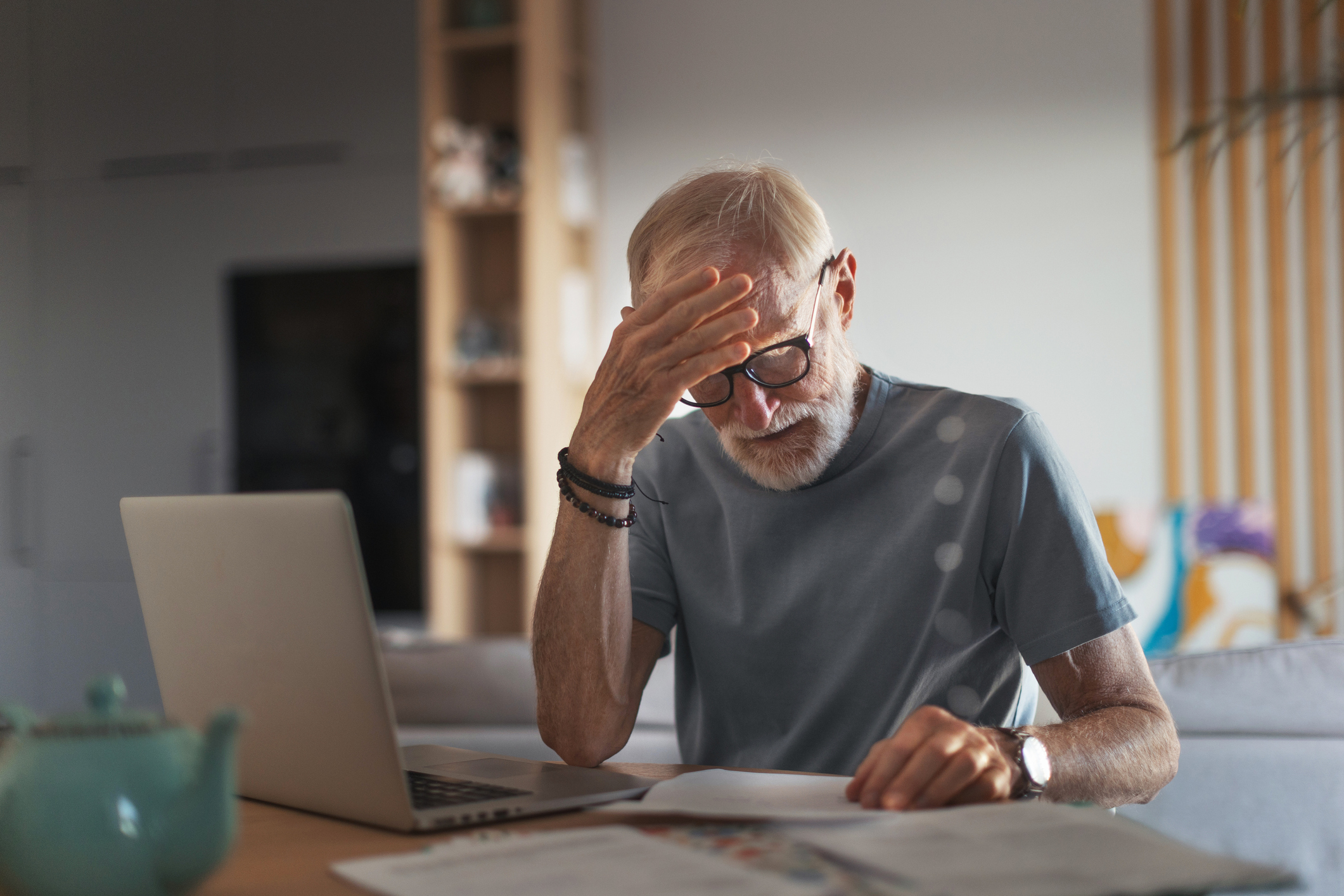 Senior man reading papers while working at his laptop in his home.