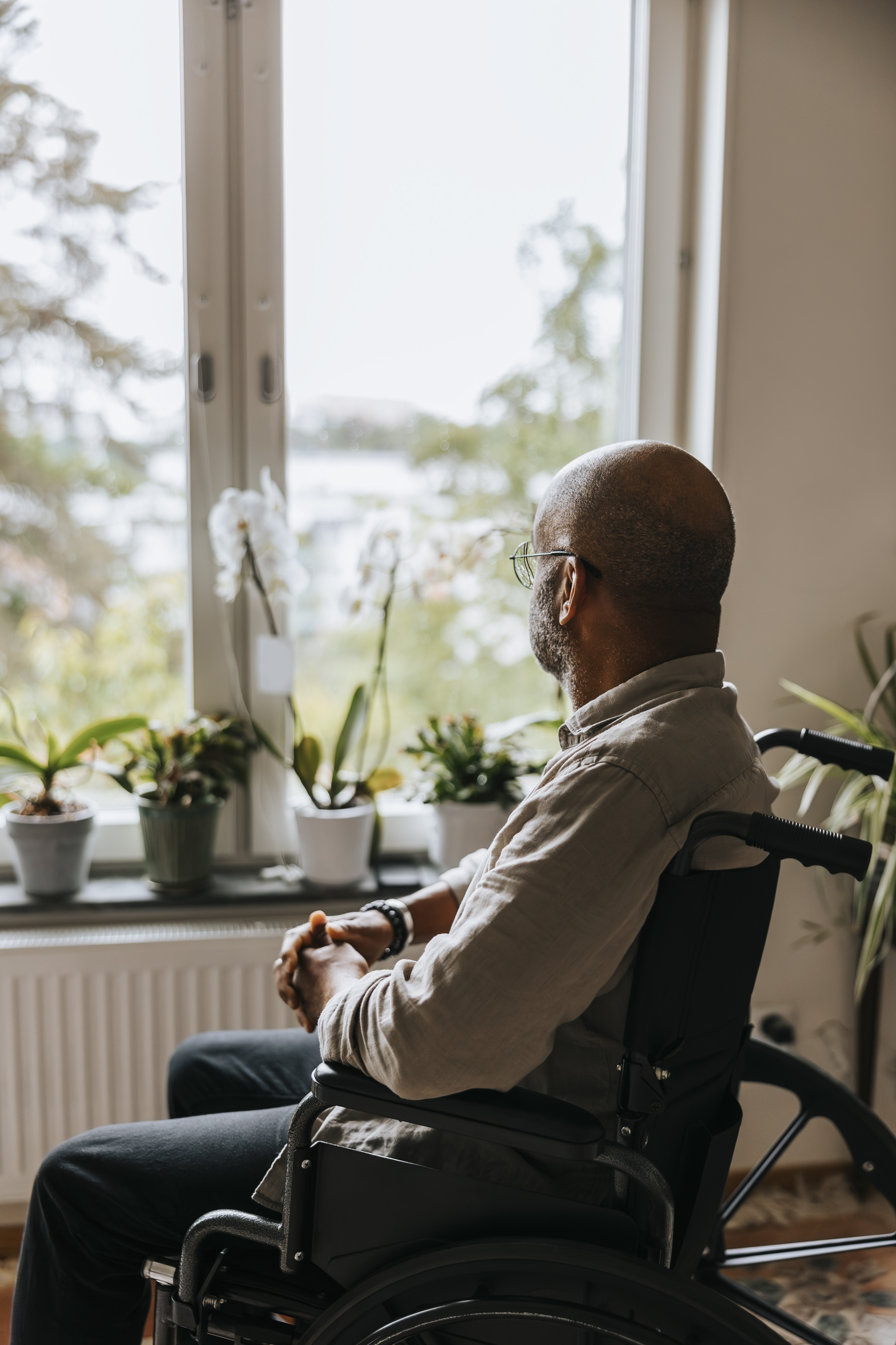 Lonely elderly man looking through window while sitting in wheelchair at nursing home