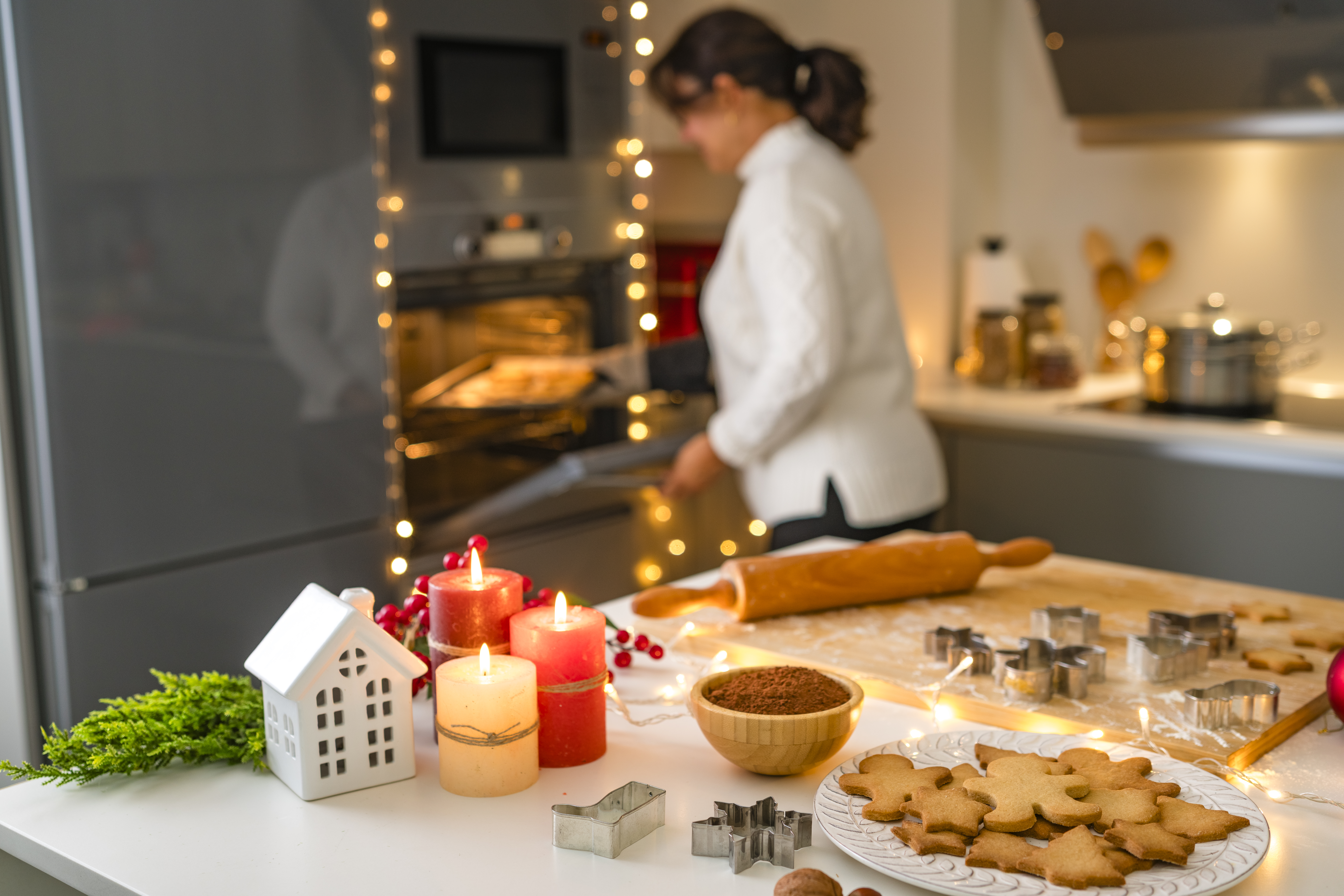 Woman placing a baking sheet full of homemade gingerbread cookies in the oven. Ingredients and utensils for preparing sweet food for Christmas Holidays celebration.