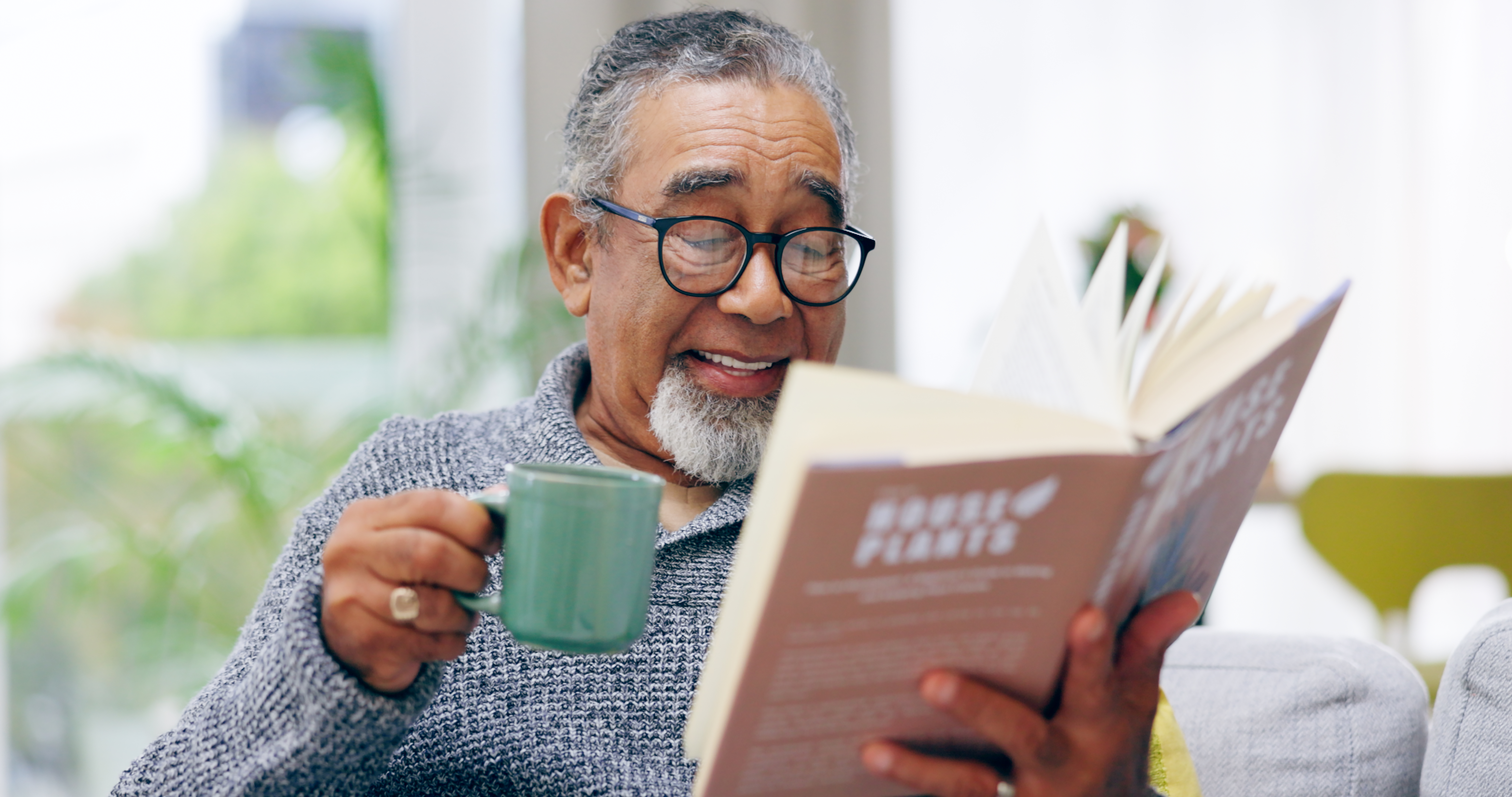 Elderly man, book and coffee on sofa with smile, reading or relax in retirement in home living room. Senior african person, literature and happy with tea for knowledge, thinking and drink on couch