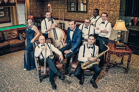 Photo of a woman in a long blue dress and 7 male band members in trousers, white shirts and suspenders with their musical instruments.