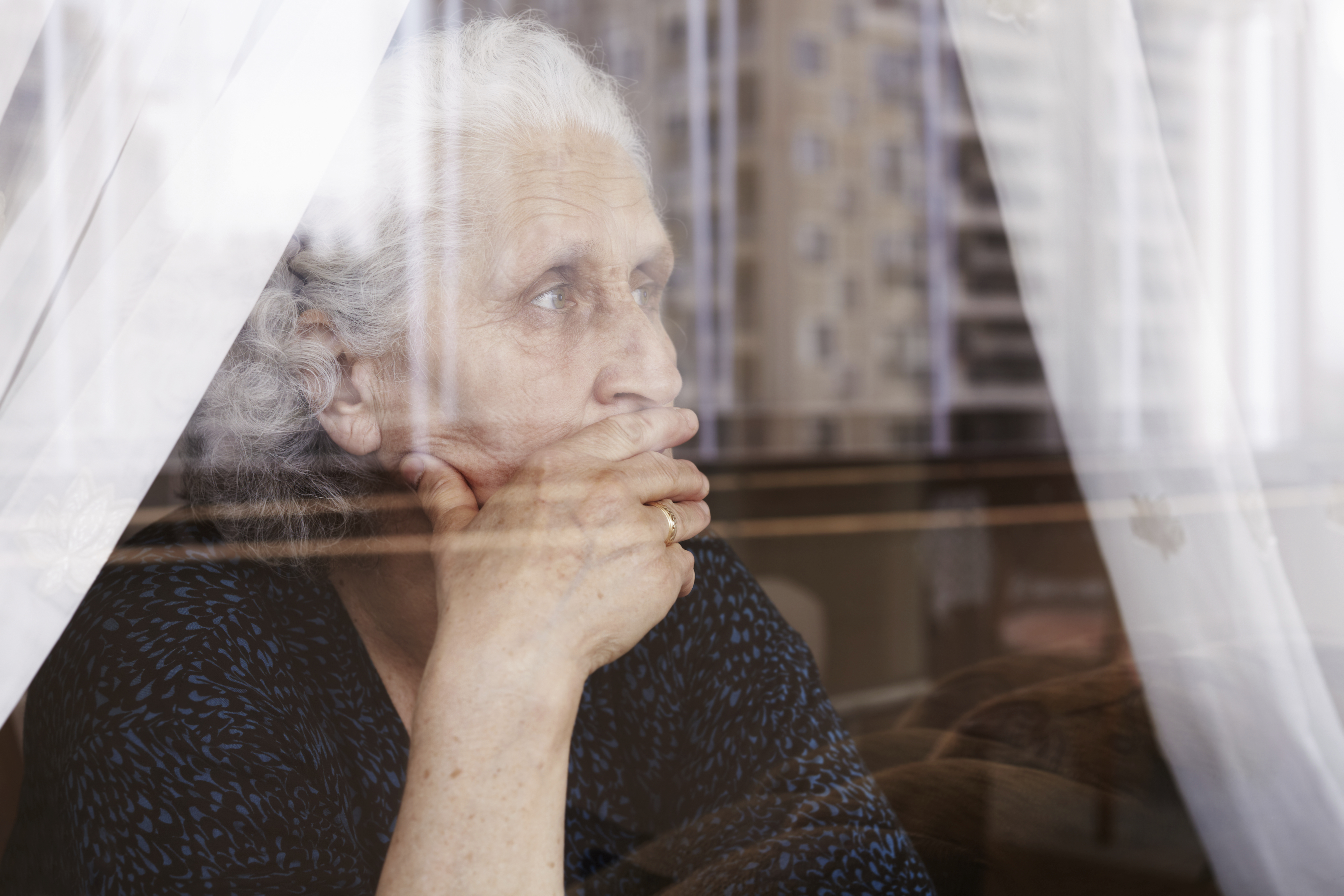 Older woman looking out a window. She appears stressed, with a hand over her mouth. 