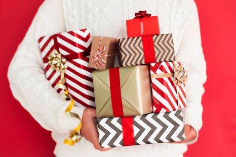 A person in a white sweater holding colorful holiday packages in front of a red packground.