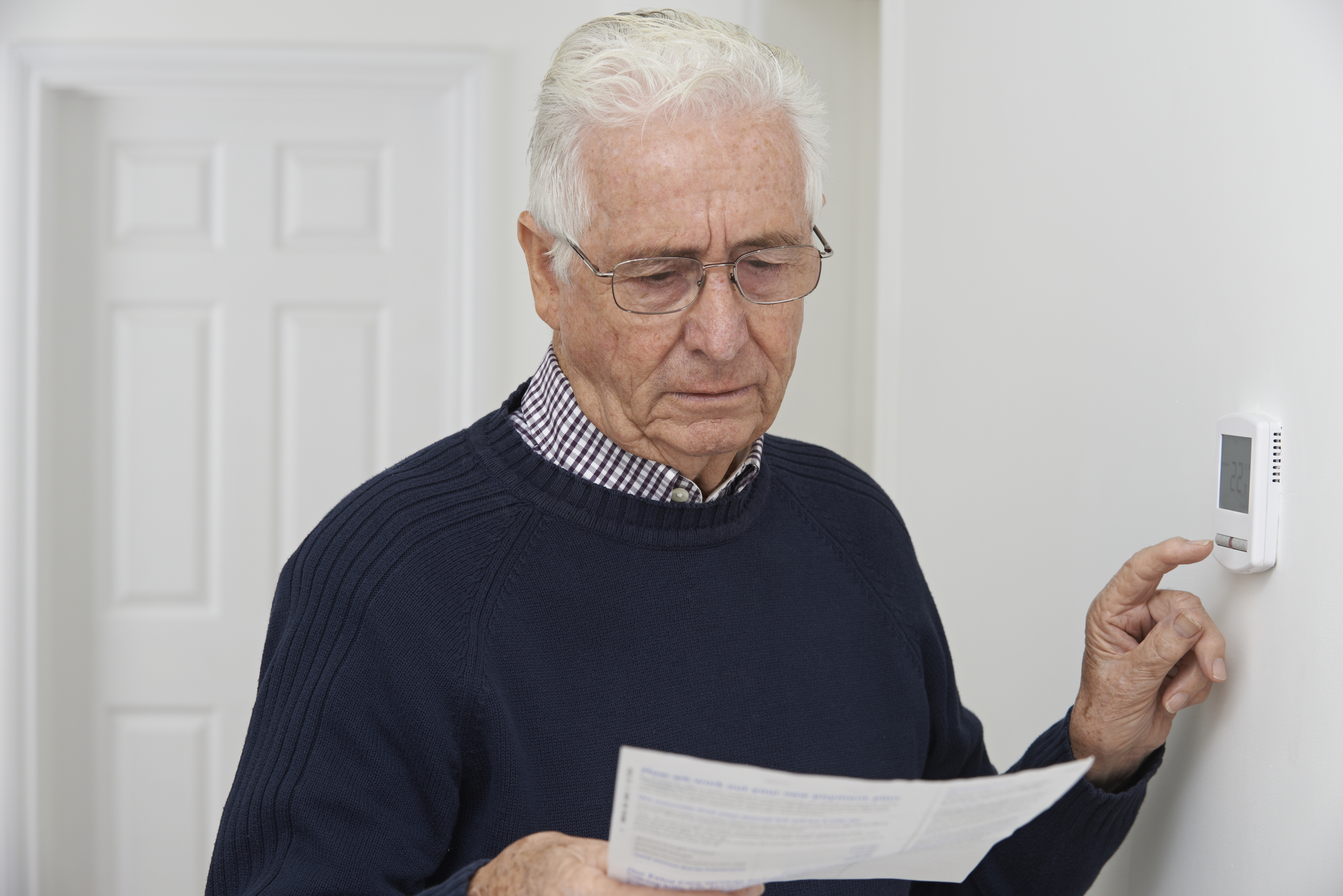 Worried Senior Man With Bill Turning Down Central Heating Thermo