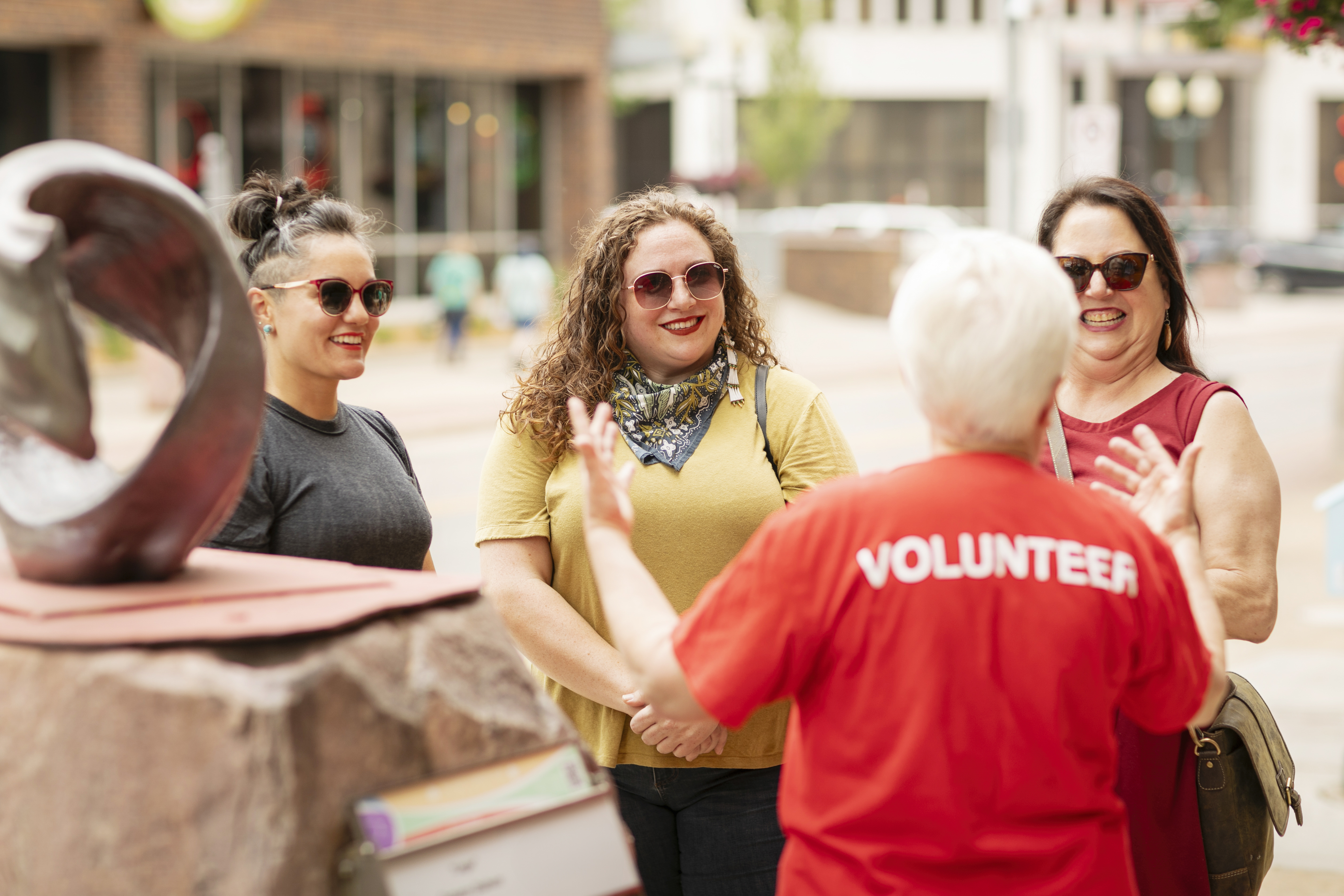 A volunteer in a red shirt explaining artwork to three female pedestrians in a downtown area.