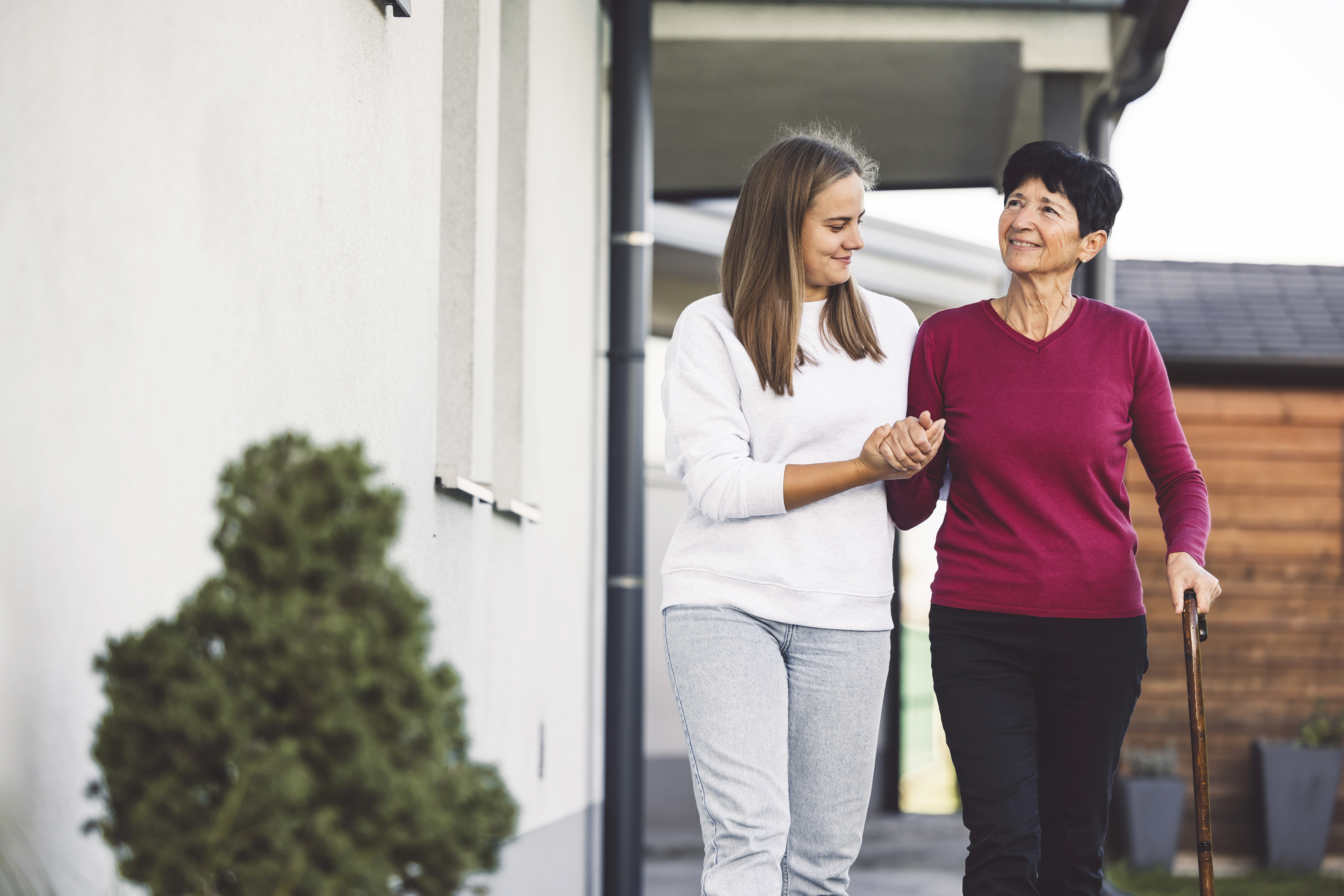 A caregiver helps an elderly woman walk in front of her house
