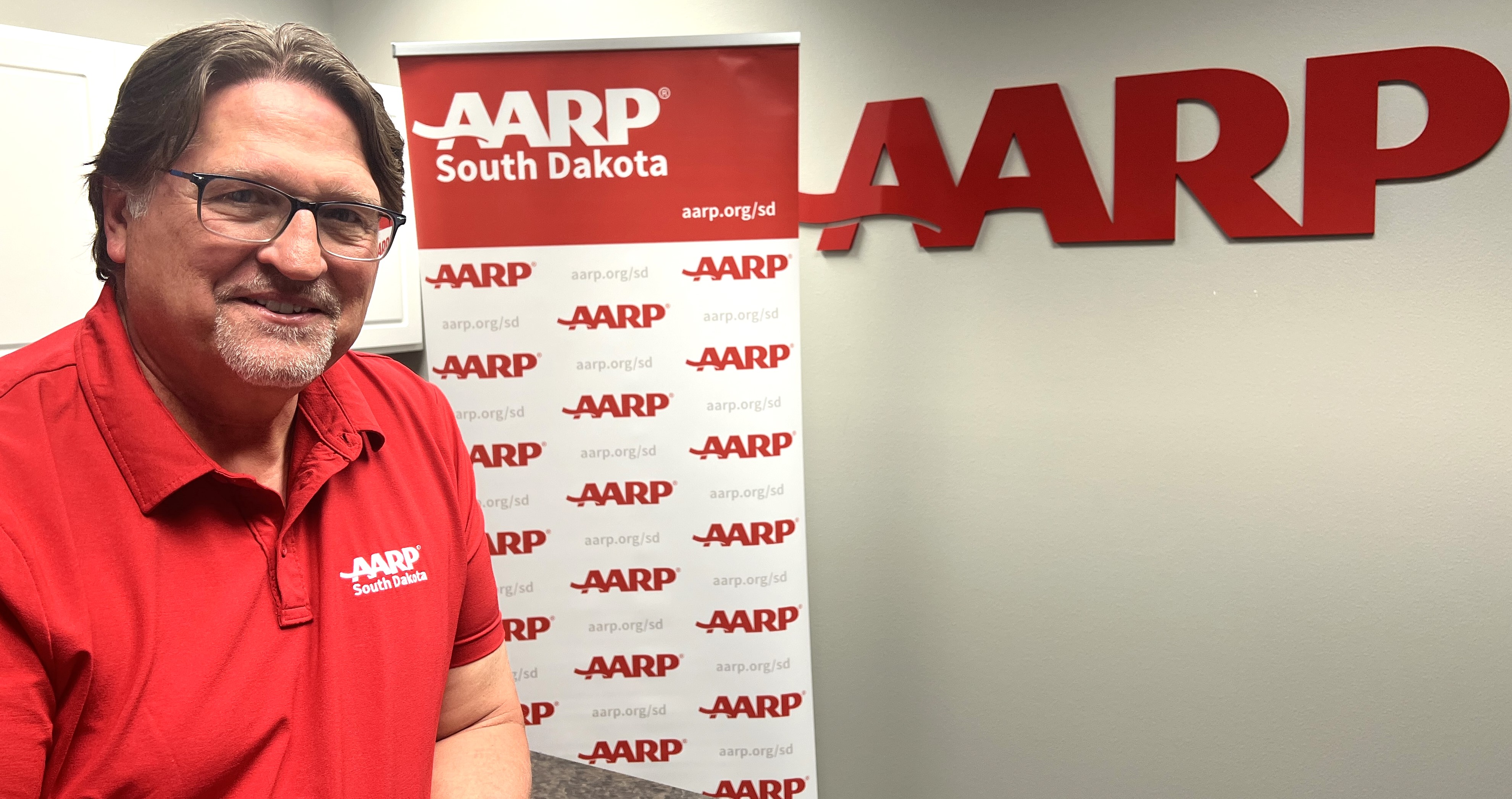 A picture of Doug DeGroot wearing a red AARP South Dakota polo shirt standing in front of an AARP South Dakota sign.