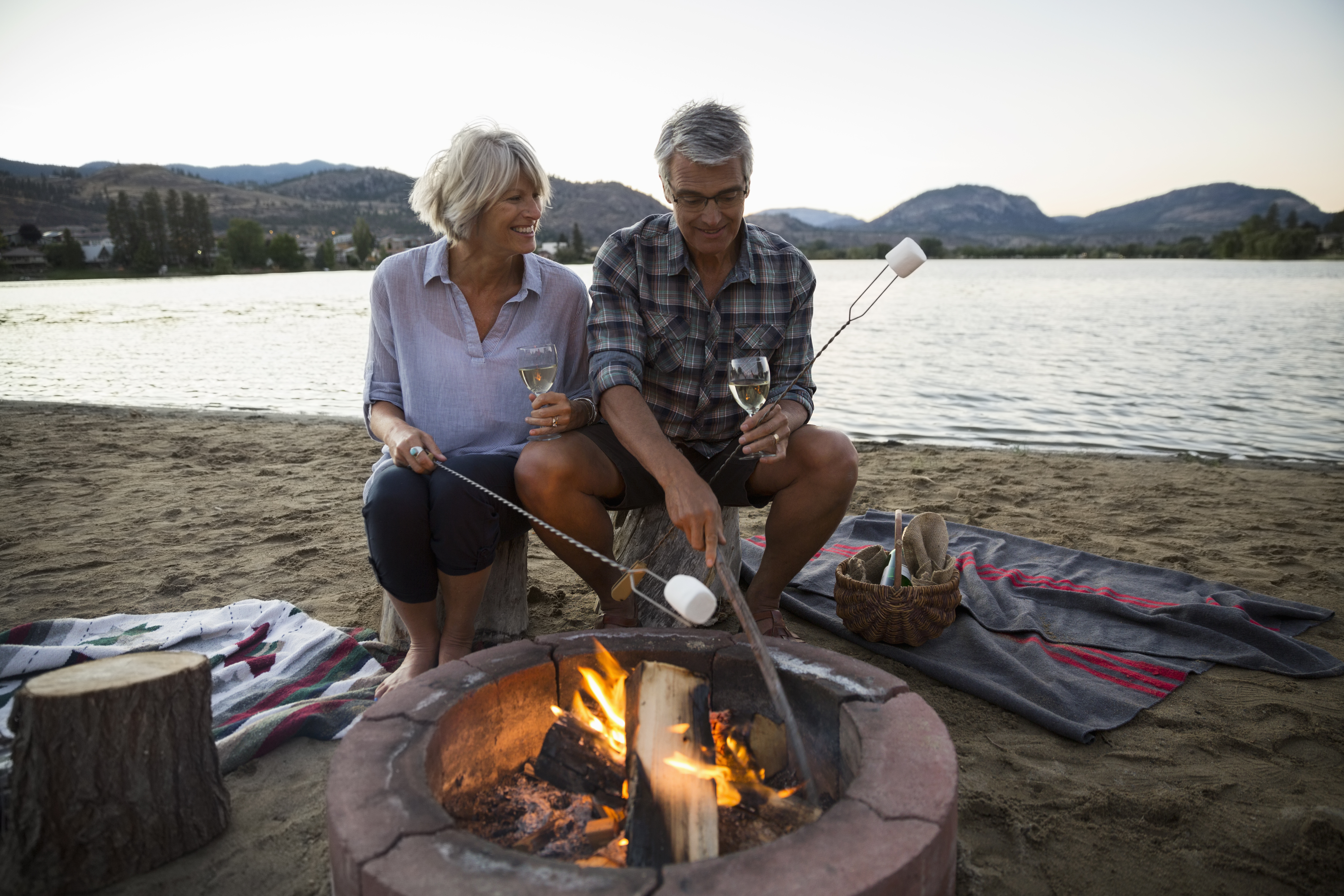 Retired couple roasting marshmallows and drinking white wine at fire pit on summer lake beach