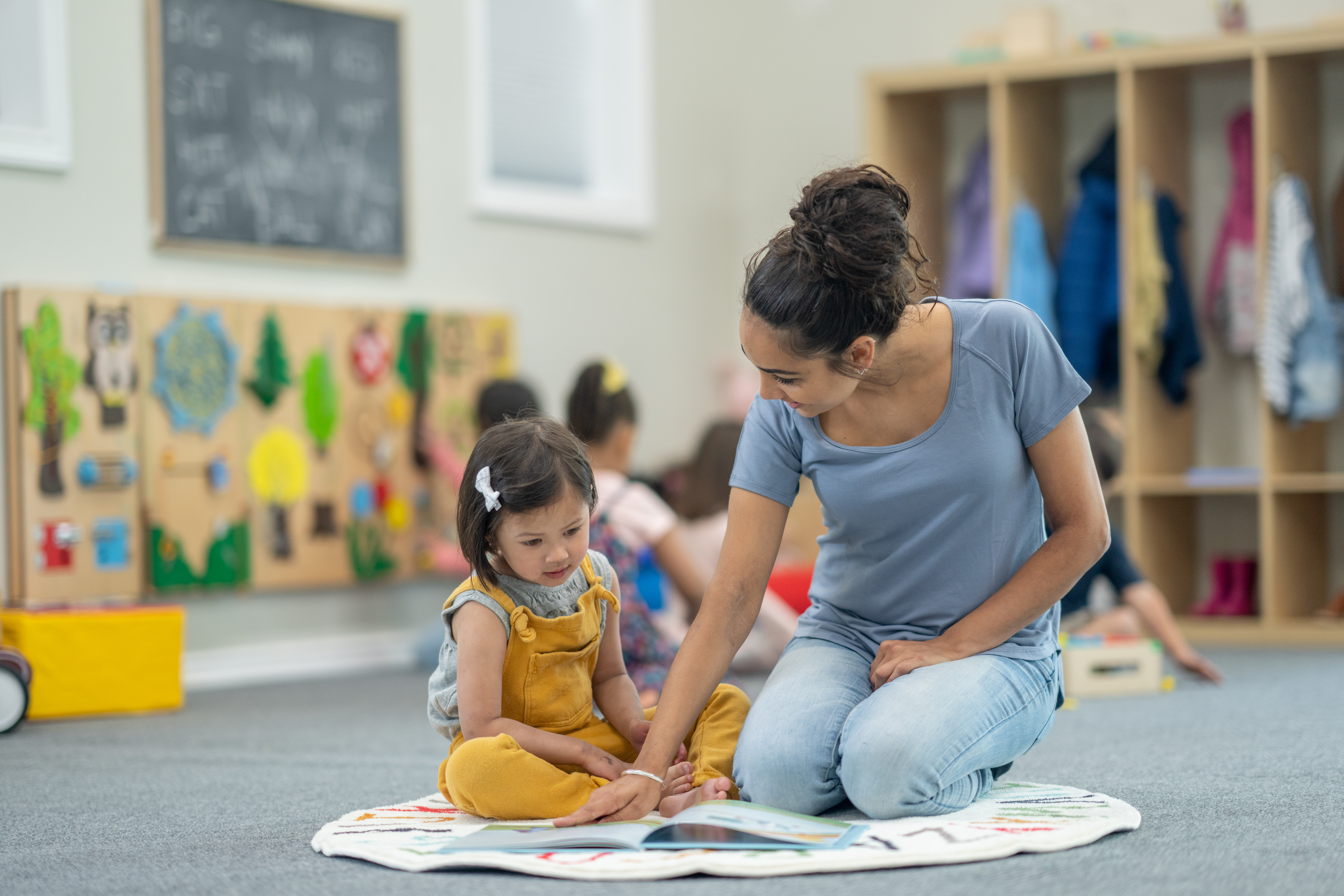 A young girl of Asian decent sits with her female kindergarten teacher as they read a book together on the rug. They are both dressed casually and the little girl is paying close attention to the story.