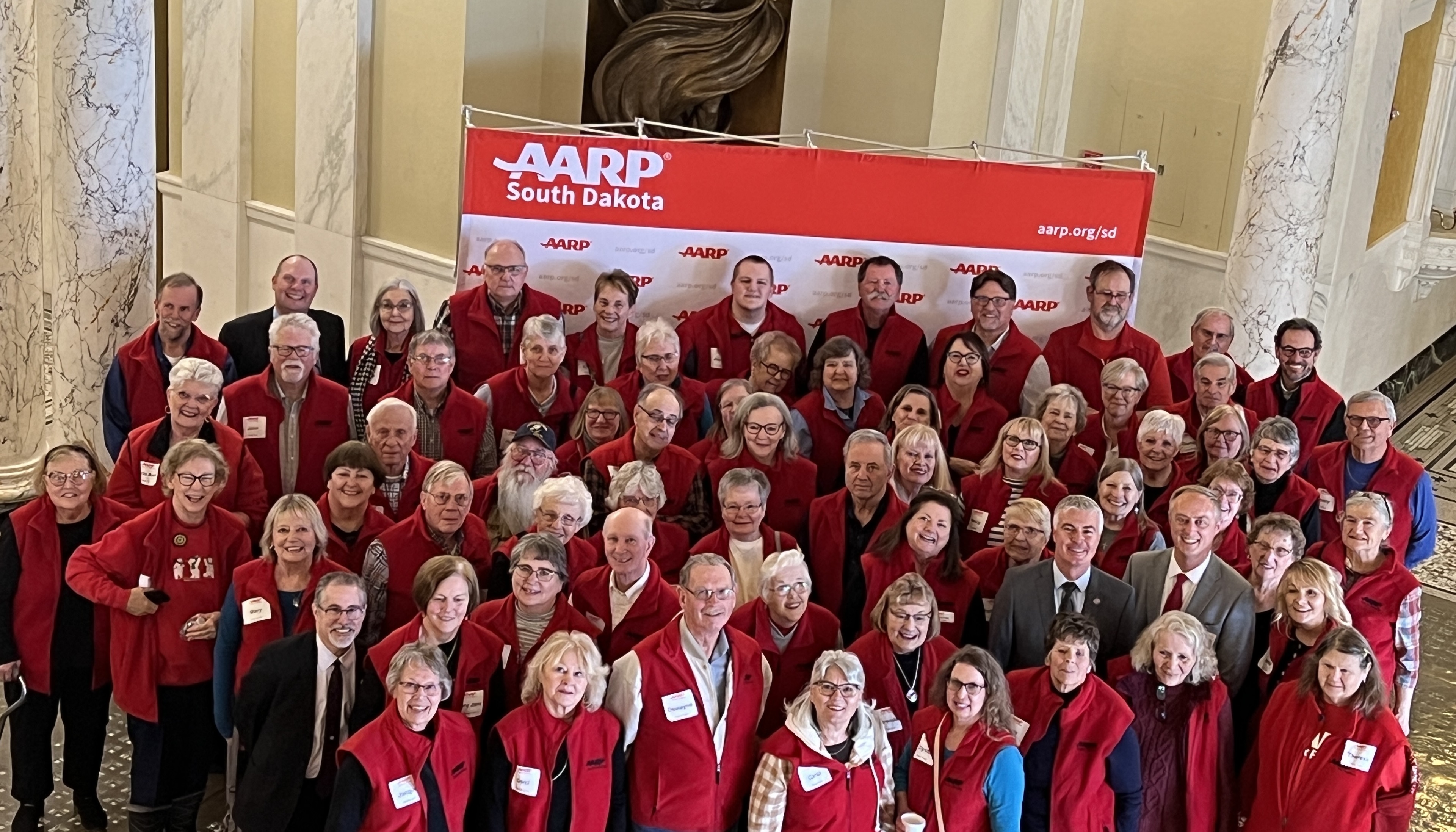 Group of 70 men and women in red AARP South Dakota vests gathered for a photo in the South Dakota Capitol Rotunda.