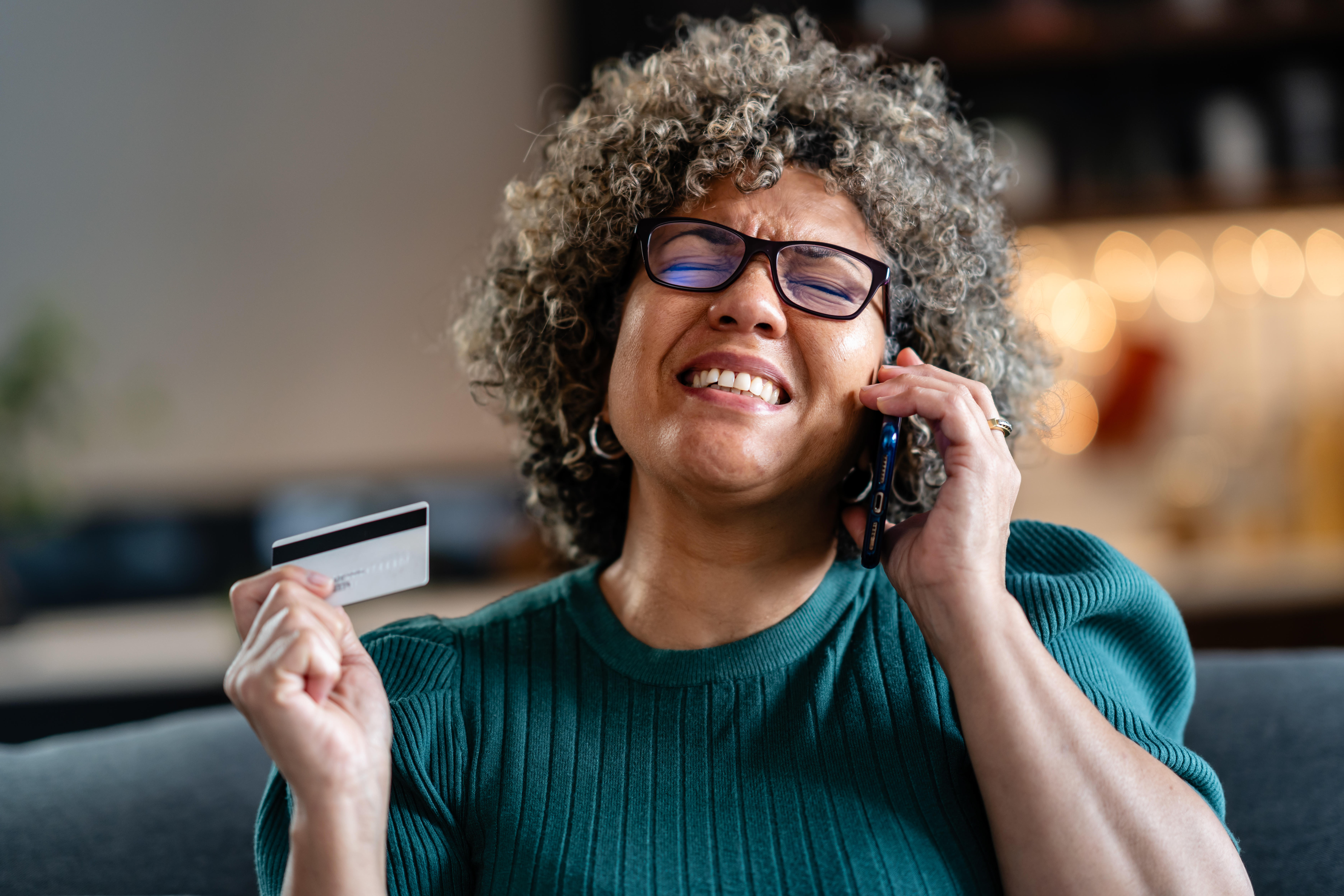 A shocked woman with curly hair and glasses is holding a credit card while talking on her phone, looking concerned and confused, possibly dealing with an issue or fraud.