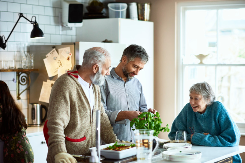 Adult son preparing meal for parents in kitchen with mother watching and smiling