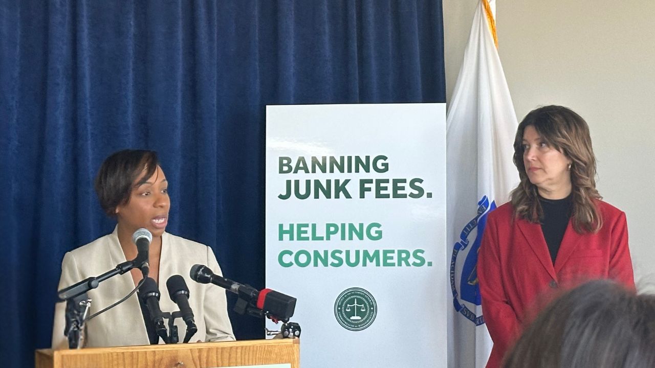 Massachusetts Attorney General Andrea Campbell standing by a sign that reads banning junk fees helping consumers. AARP Massachusetts State Director Jen Benson is standing by, waiting to speak. 