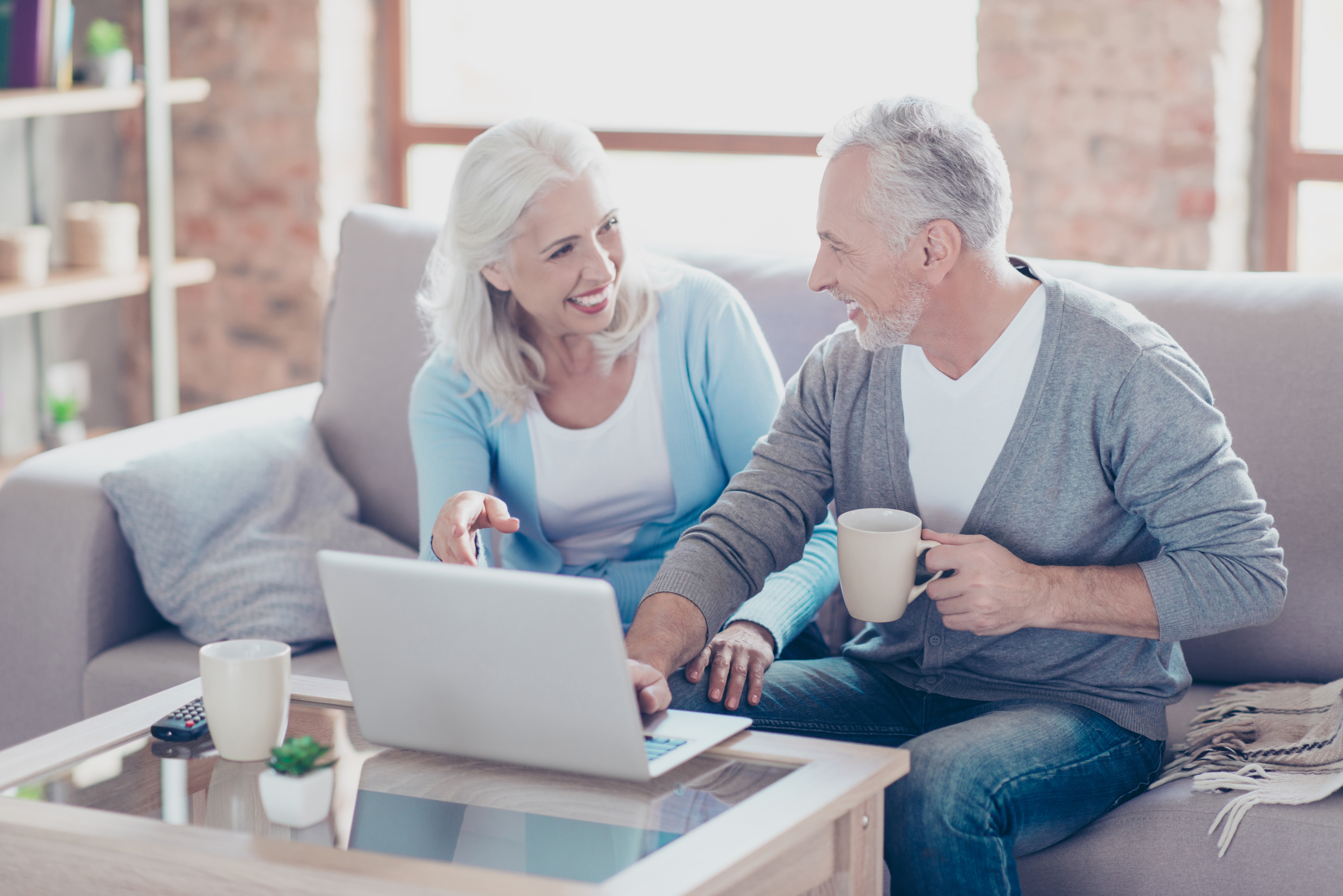 Two elderly people are sitting at home at the weekend, drinking coffee and watching films using the computer on the table, the woman is pointing on the monitor