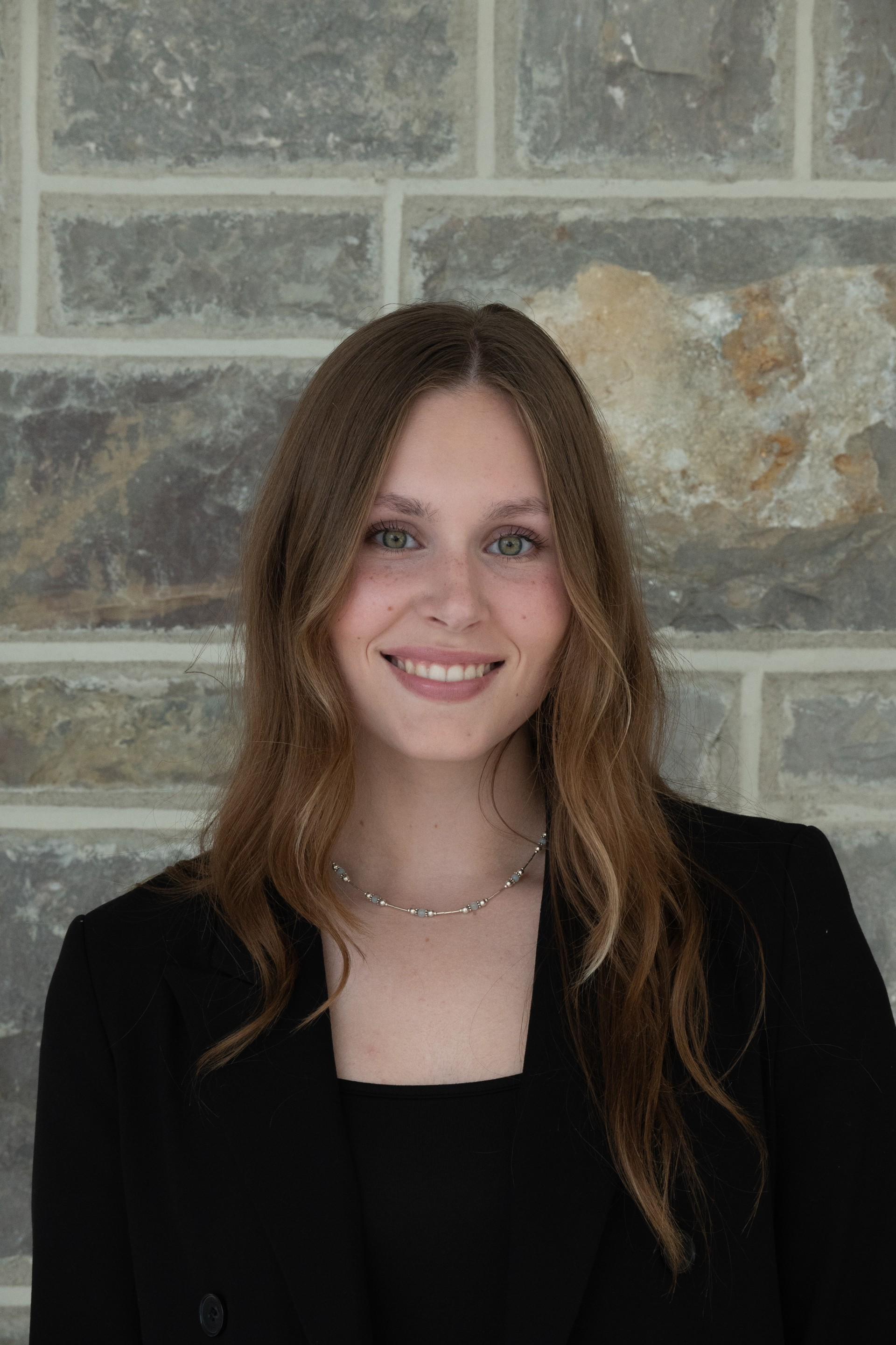 A young woman in a black blazer poses in front of a stone wall.