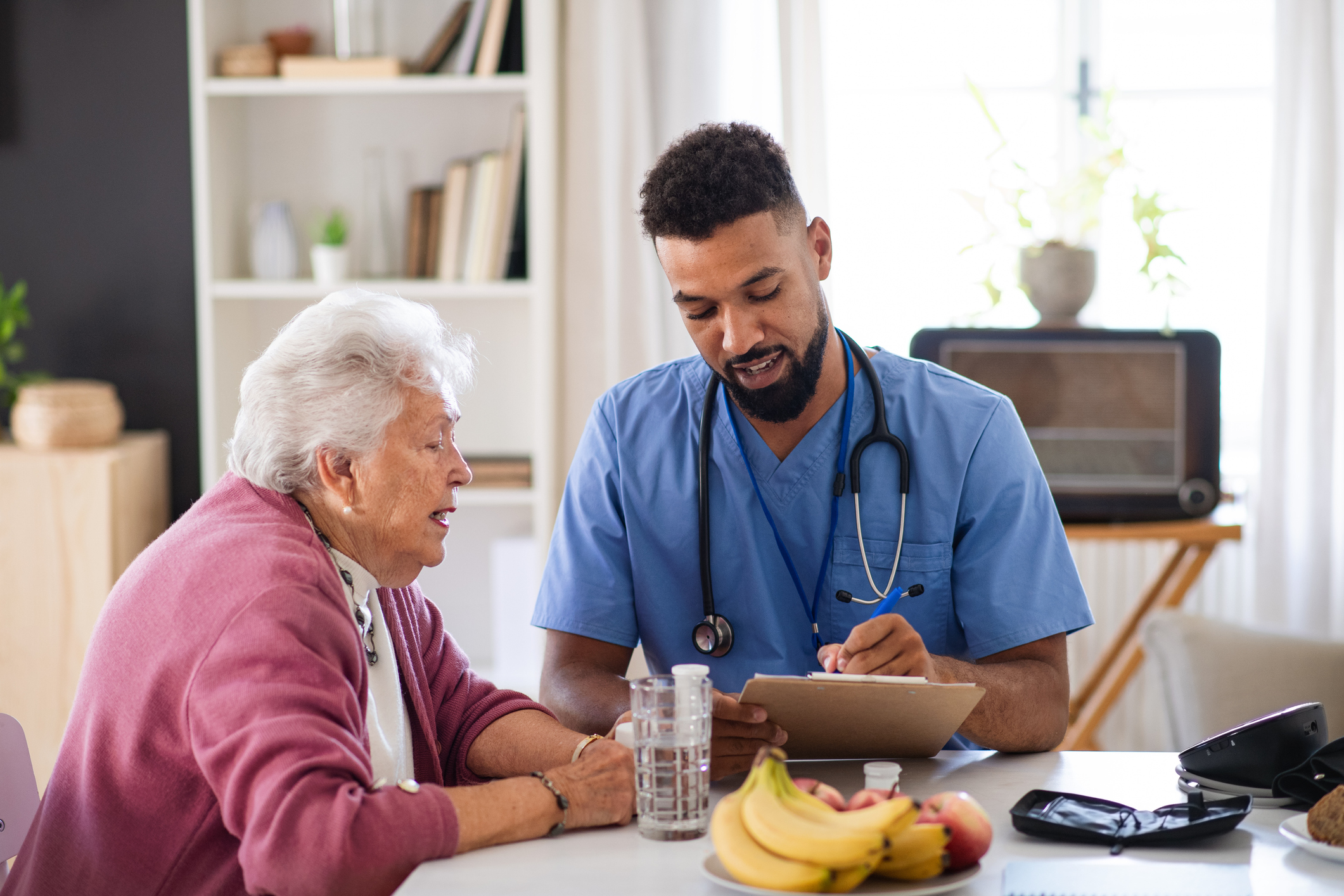 Young healthcare worker or caregiver visiting senior woman indoors at home, talking and writing.