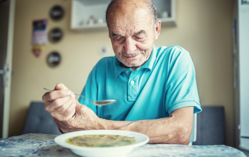 Elderly man holds his hand trying to control Parkinson disease while eating a soup.