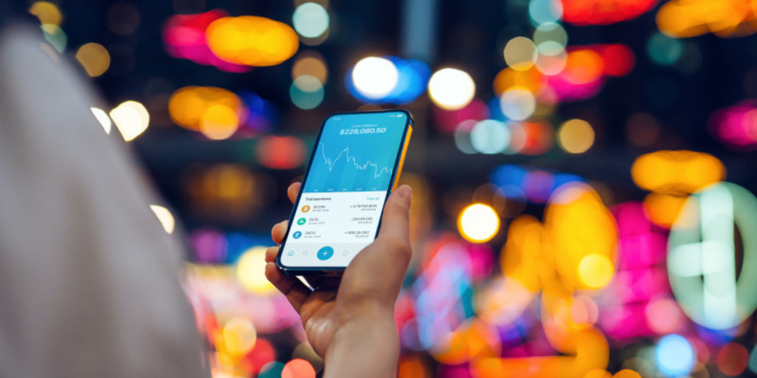 A person holds a mobile phone and looks at their crypto account with colorful lights out of focus in the background.