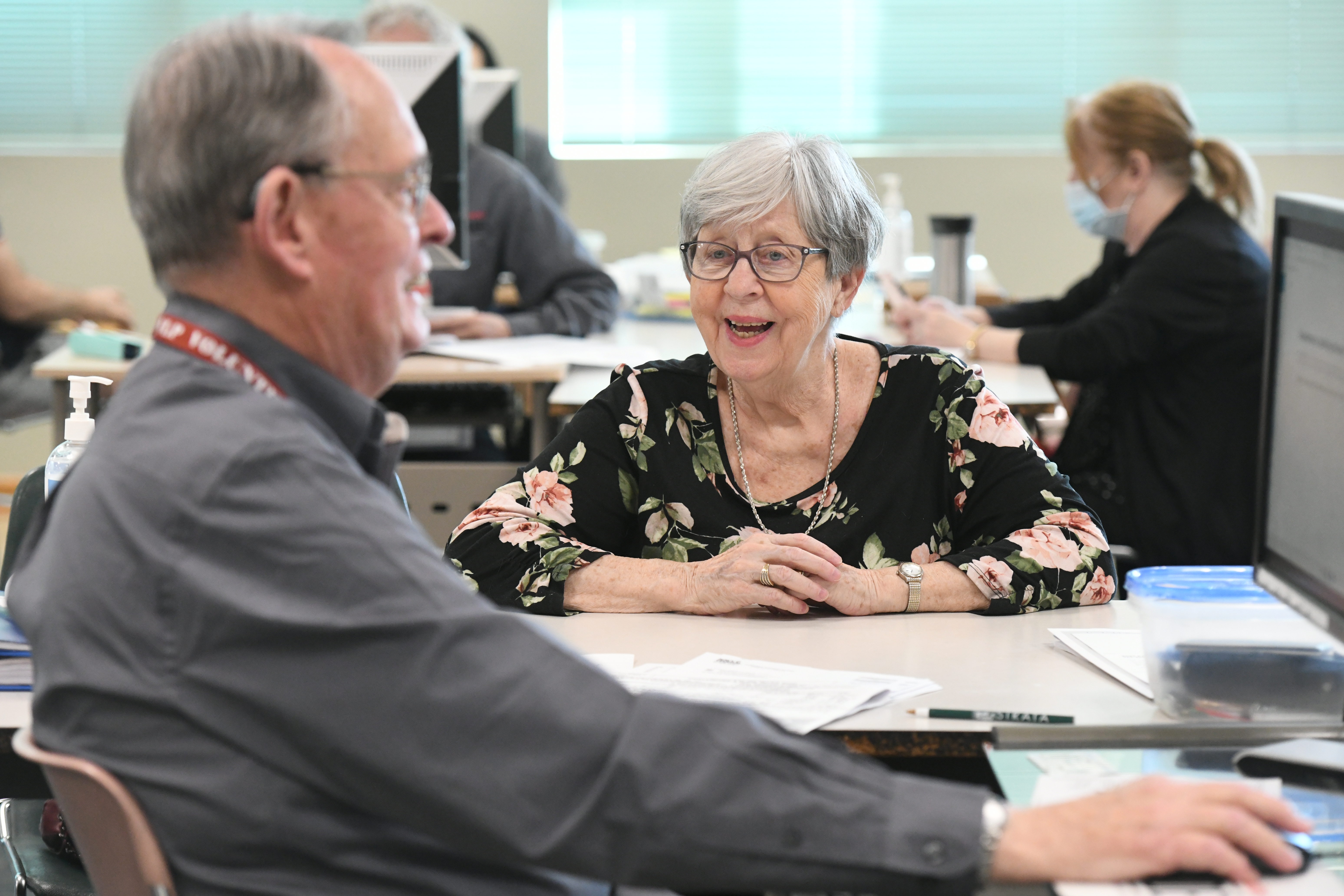 Middle aged man in a grey shirt is a tax preparation volunteer helping an older woman fill out her tax forms.