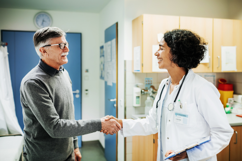 An older male patient shakes hands with a female health care provider in a white lab coat.