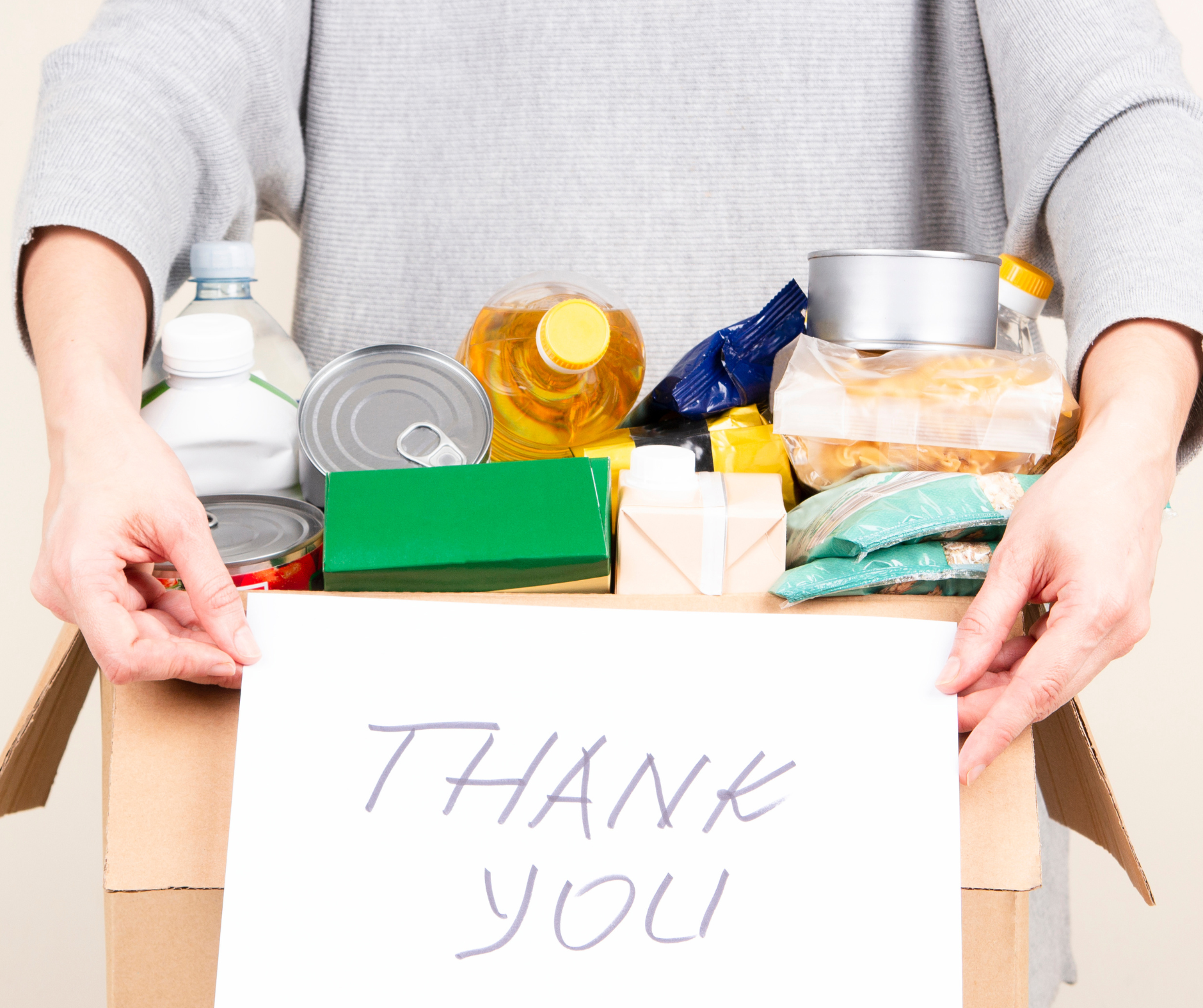 Women's hands holding a note that says thank you in front of a box of donated items for a food pantry.