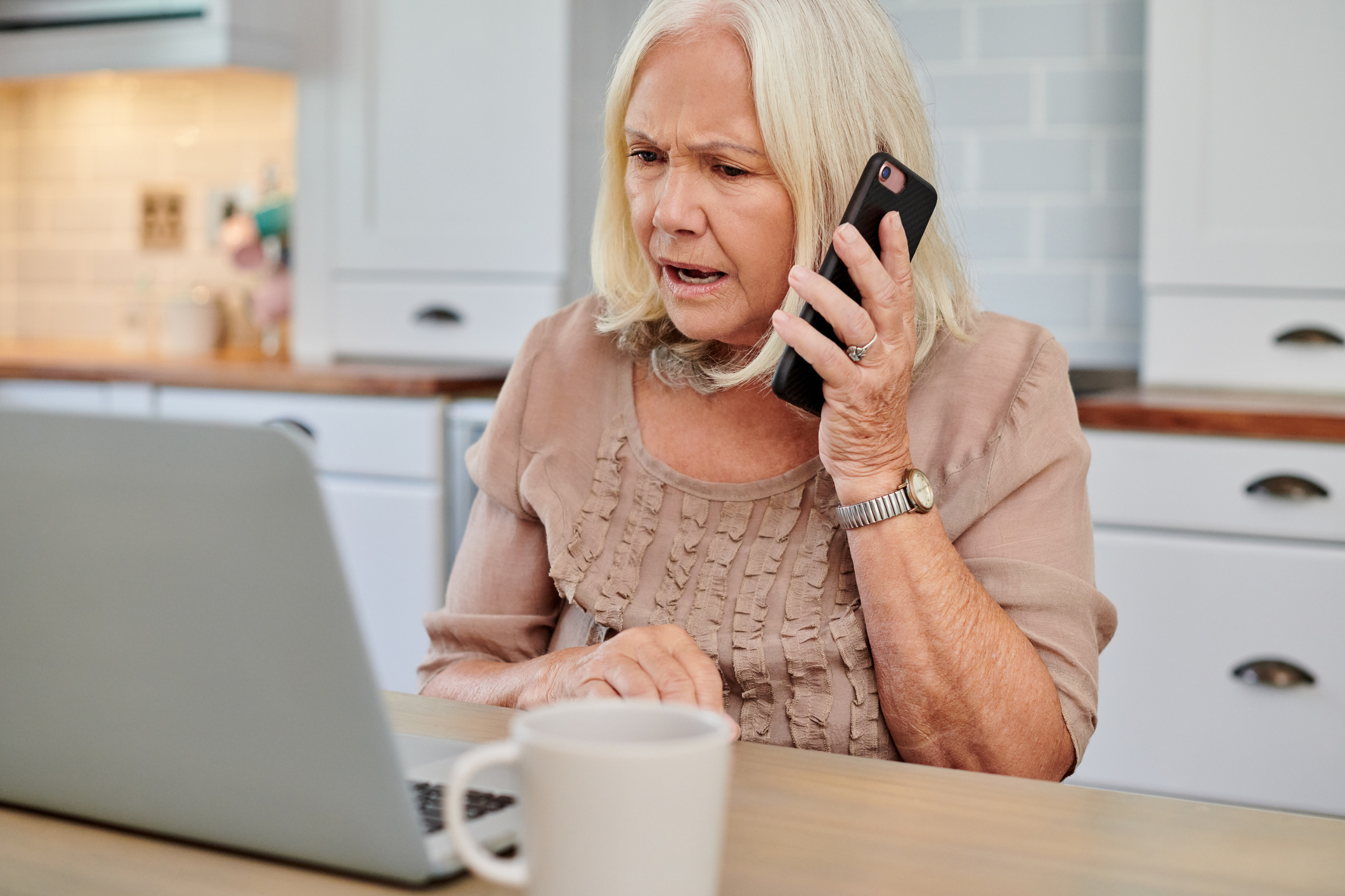 Shot of an older woman looking upset while using a smartphone and laptop at home.