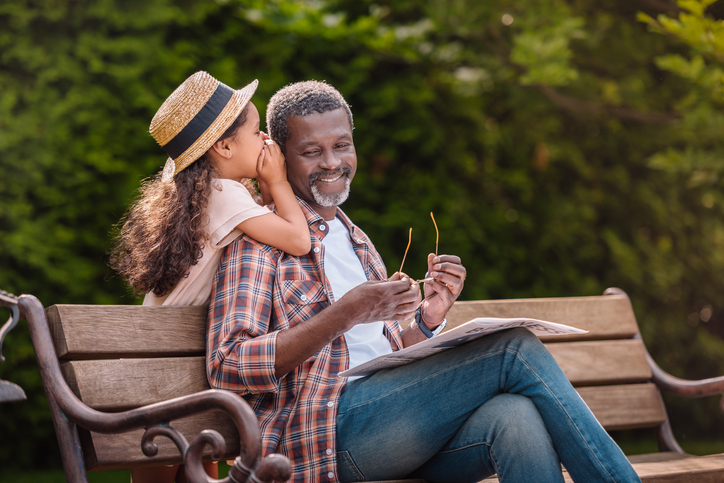 grandchild whispering to her smiling grandfather while sitting on bench in park