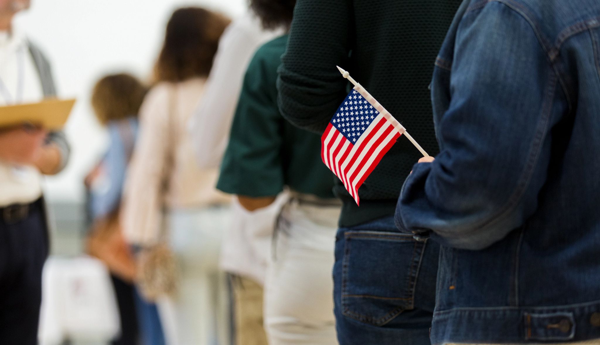 A diverse group of unrecognizable people stand in line to vote. A woman at the end of the line holds an American flag.