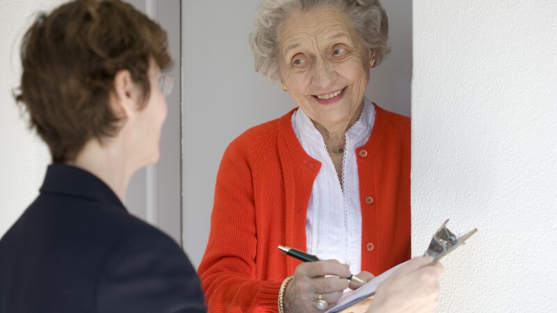 Smiling senior woman at her door signing a document on a clipboard held by a woman