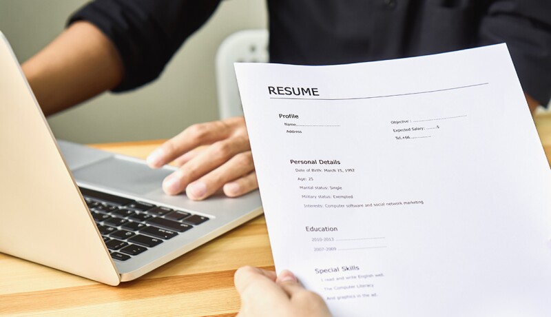 Cropped Image Of Person Holding Resume Against Business Person Using Laptop At Table