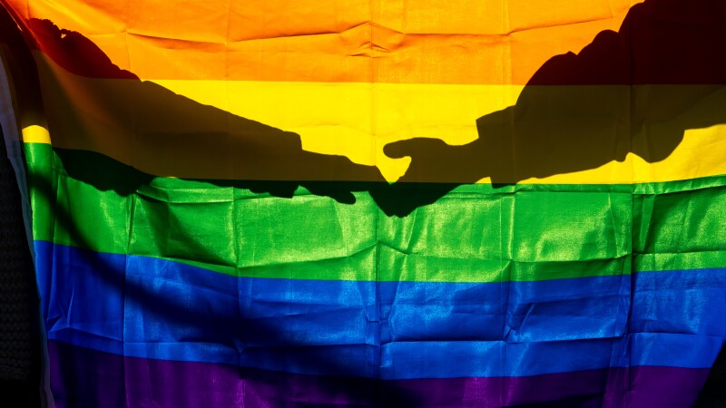 Two women are holding hands against the light through the LGTBI flag.