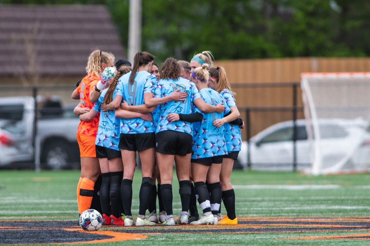 A group of female soccer players in blue jerseys in a team huddle.