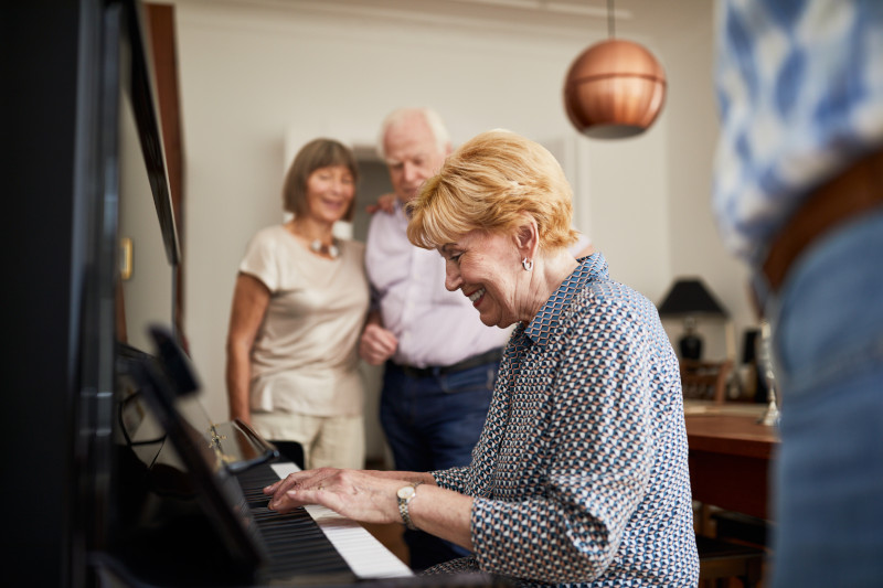 Smiling elderly woman playing the piano with friends listening