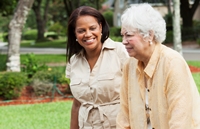 Caregiver (50s) helping senior Hispanic woman (70s) with walker outdoors.