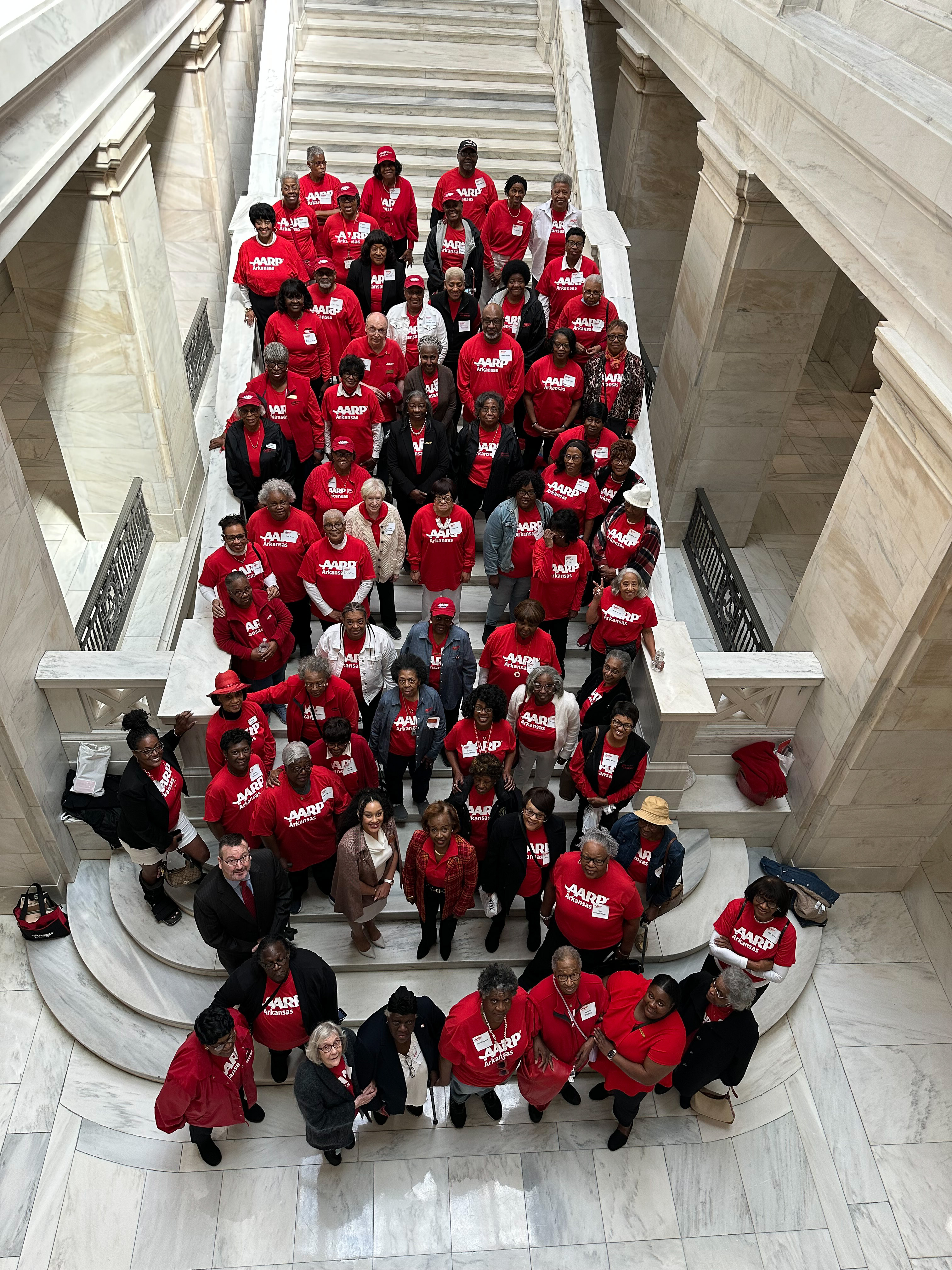 A group of volunteer stand on the steps to the Arkansas House of Representatives and look up at the camera. The volunteers are primarily wearing red shirts that say AARP Arkansas